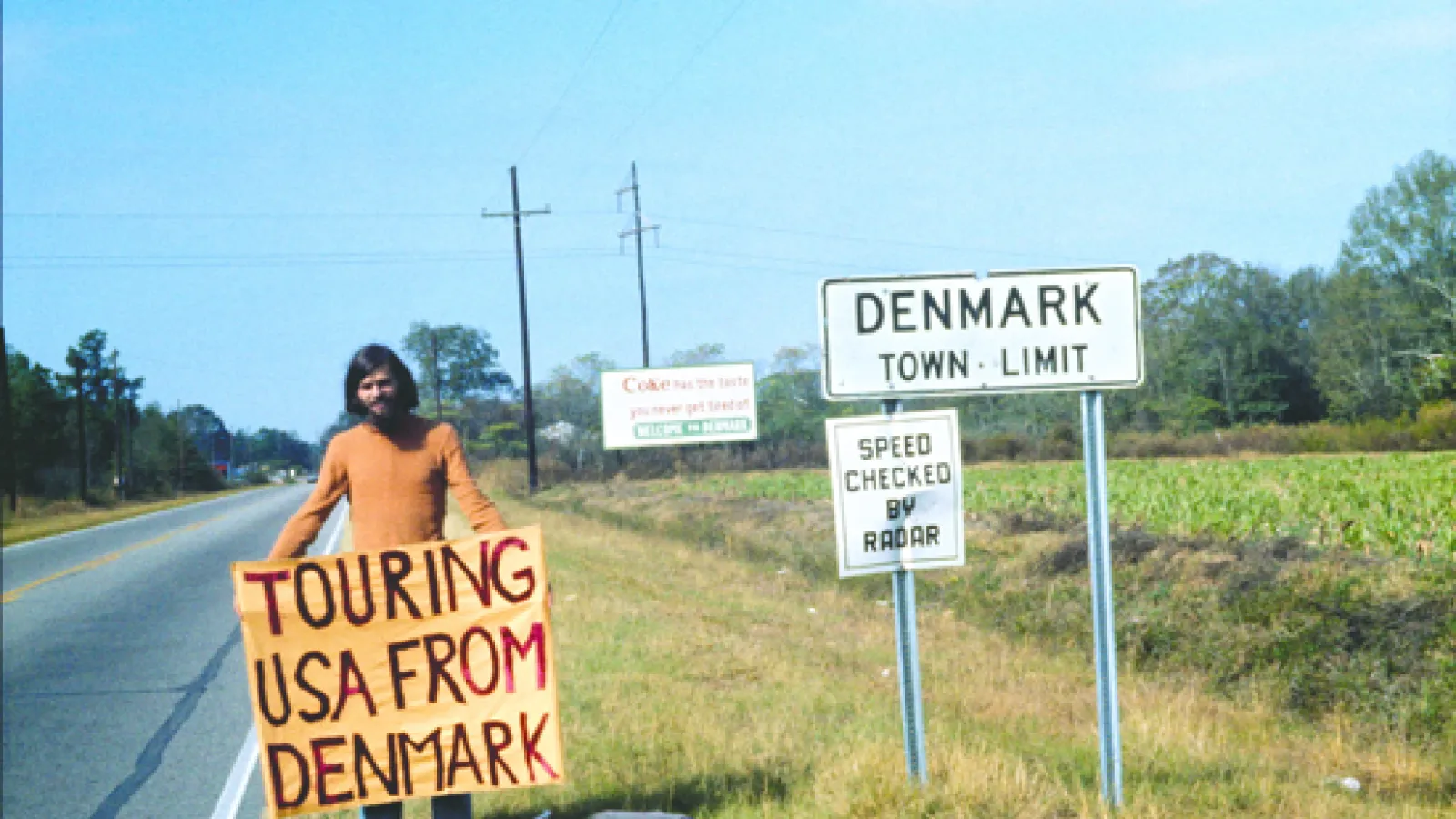 a man on the side of highway holding sign that says" Touring USA from Denmark"