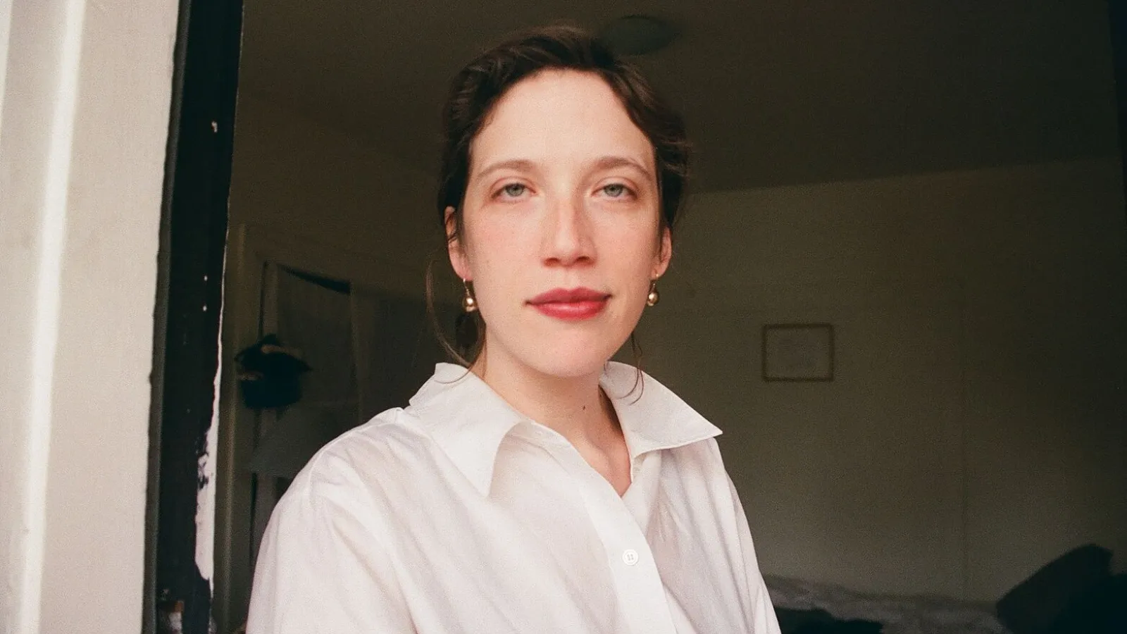 figure in white shirt with light skin, brown hair, and red lips in front of dimly lit room