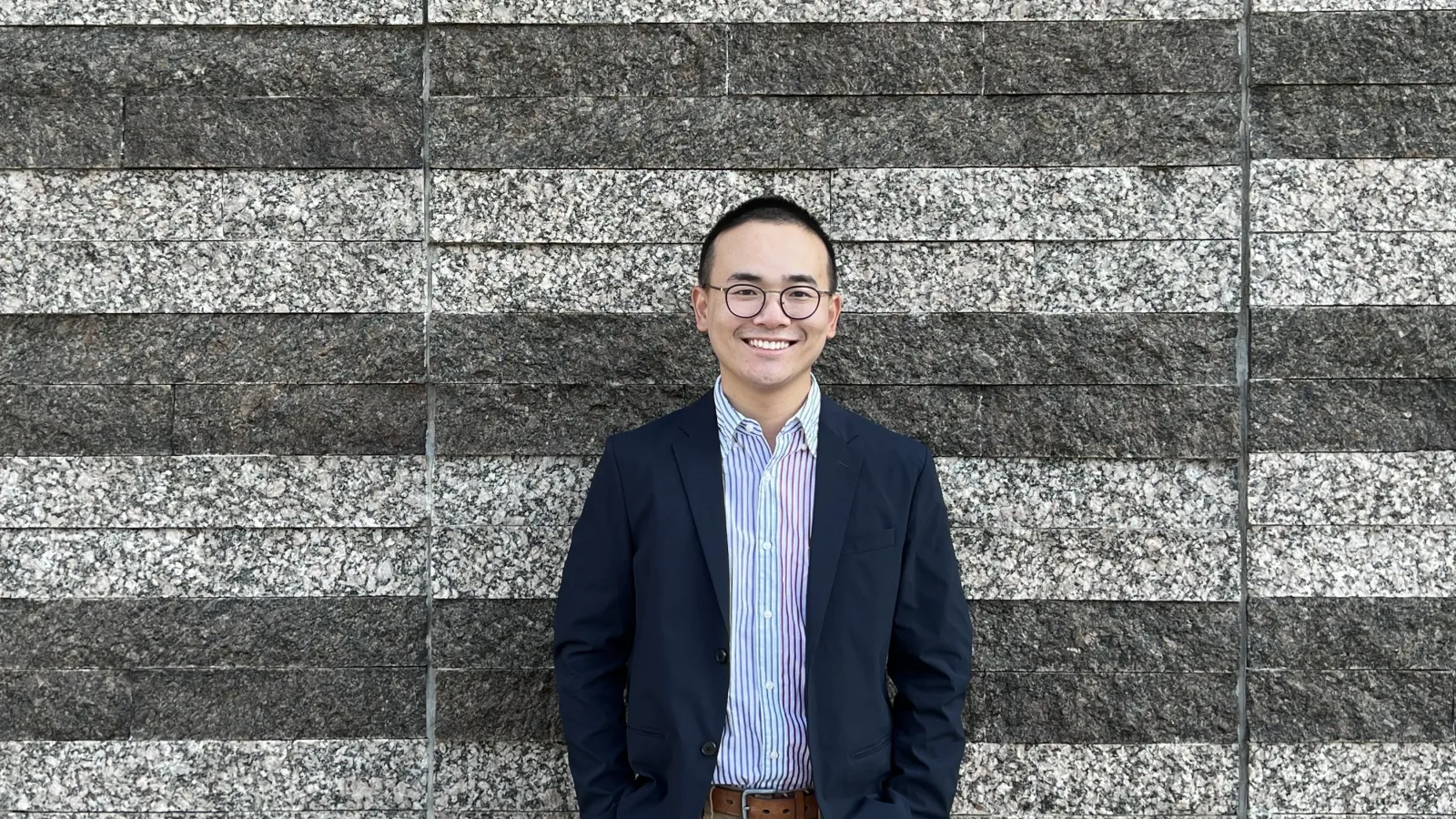 Yifan Li standing in front of a stone wall wearing a multicolor button up shirt, a navy jacket, and kaki pants.