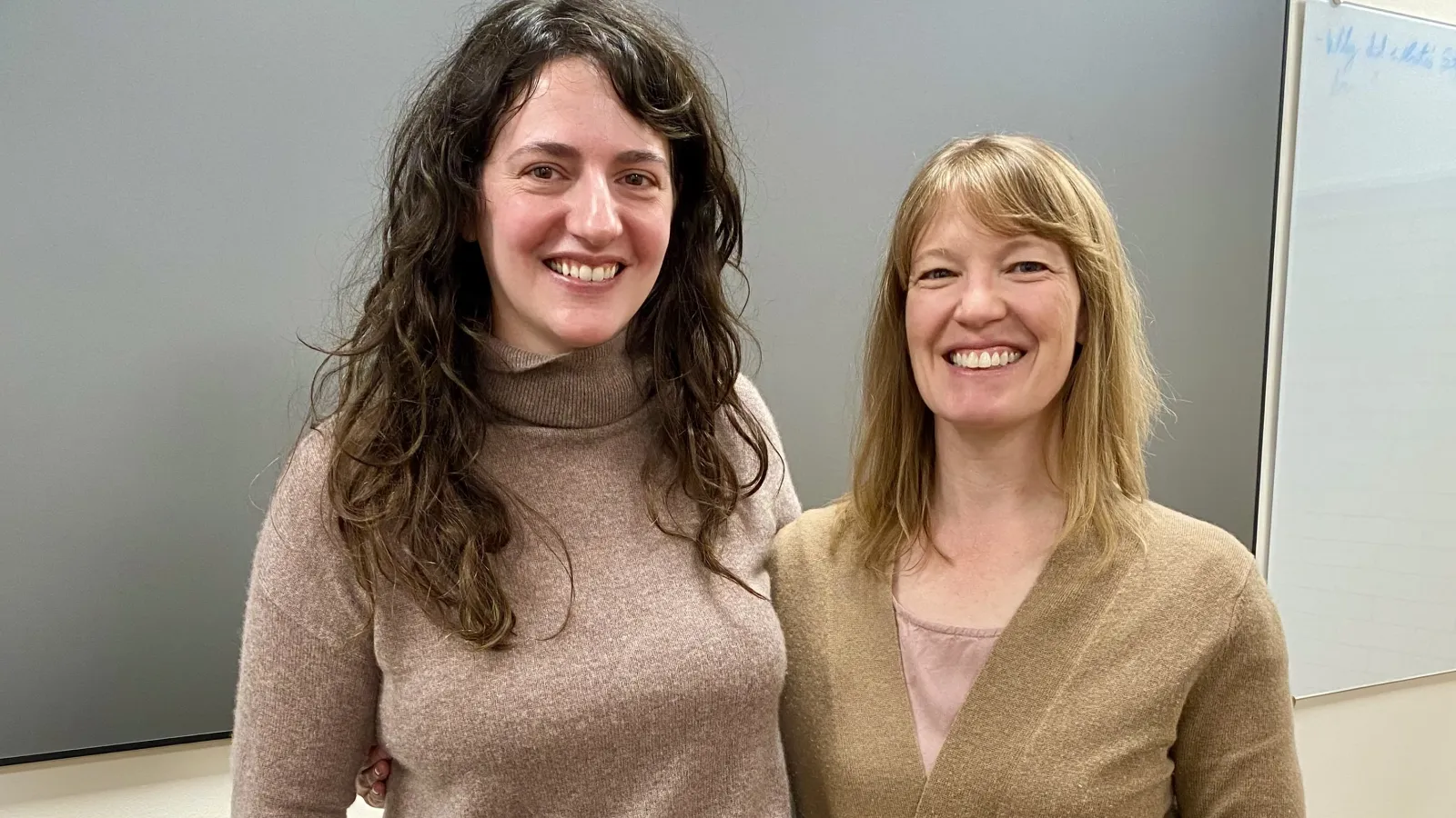Julie Defossez (left) and  Kris Paulsen (right) pose standing together both wearing beige sweaters and black pants. 