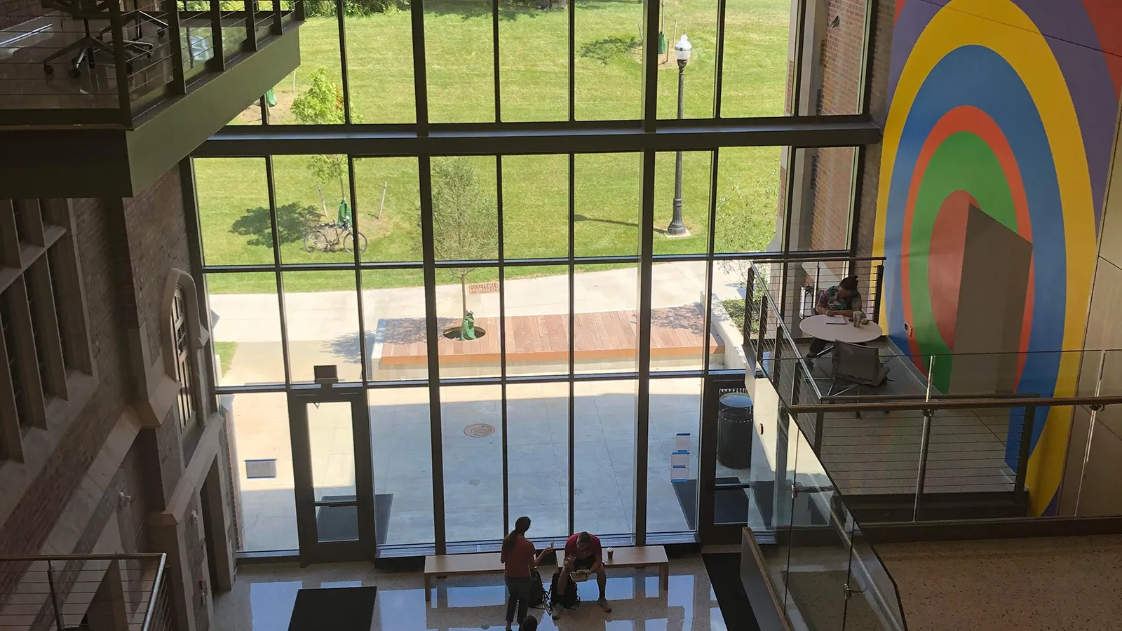 Several students sit in the interior of a building showing large windows covering the back wall, and a colorful mural by the artist Sol LeWitt on the right.