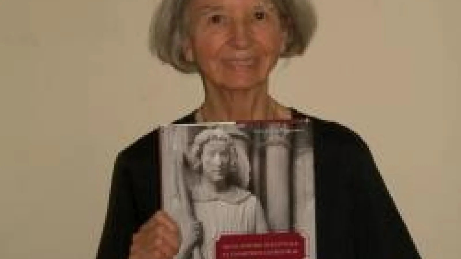  Anne Morganstern wearing black and  holding a book in front of a tan wall