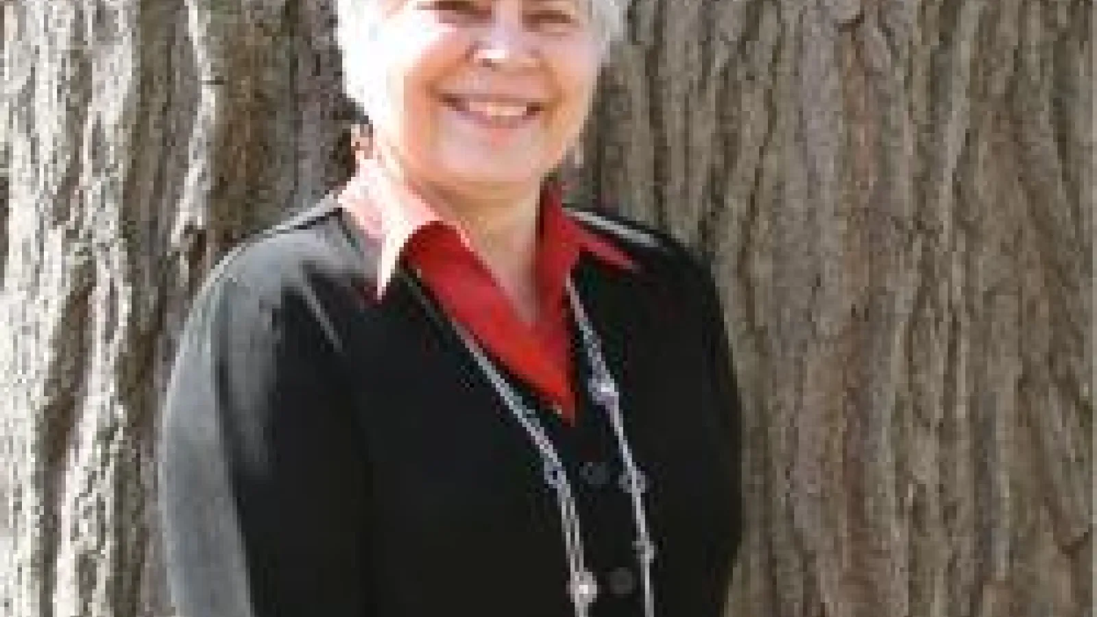 Dr. Myroslava Mudrak wearing a black blouse and red collar standing in front of a tree. 