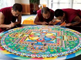 3 men in maroon outfits working on sand mandala