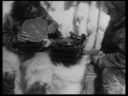 black and white film still of two figures around a record player bundled in warm clothing