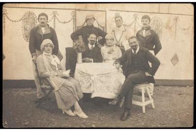 Sepia-tinted photograph of eight figures sitting around a table dressed in old-fashioned attire and looking directly toward the camera