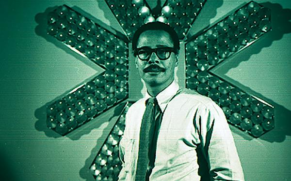 Image of man in white shirt and tie with dark glasses in front of asterisk-shaped sculpture. Phot has a green hue