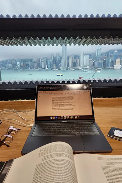 A book and an open laptop sit on a wooden surface in front of a large window, which reveals a panoramic cityscape