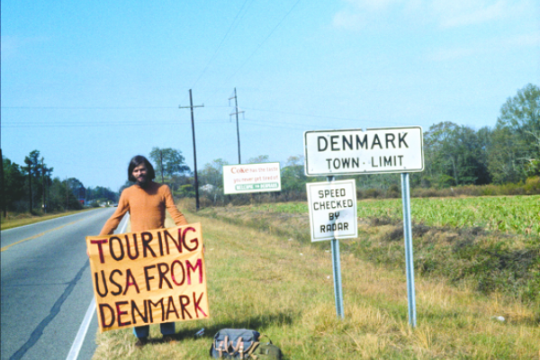 Photo of a man on the side of a highway holding a sign that says" Touring USA from Denmark"