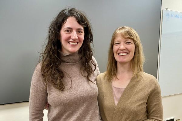 Julie Defossez (left) and  Kris Paulsen (right) pose standing together both wearing beige sweaters and black pants. 