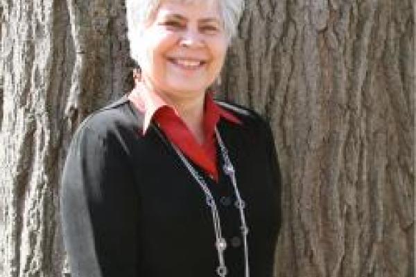 Dr. Myroslava Mudrak wearing a black blouse and red collar standing in front of a tree.