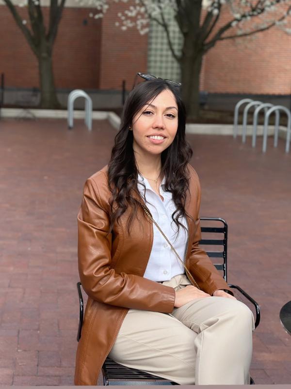 Shima Karimi sitting on a chair wearing a white top, khaki pants, and a brown leather jacket