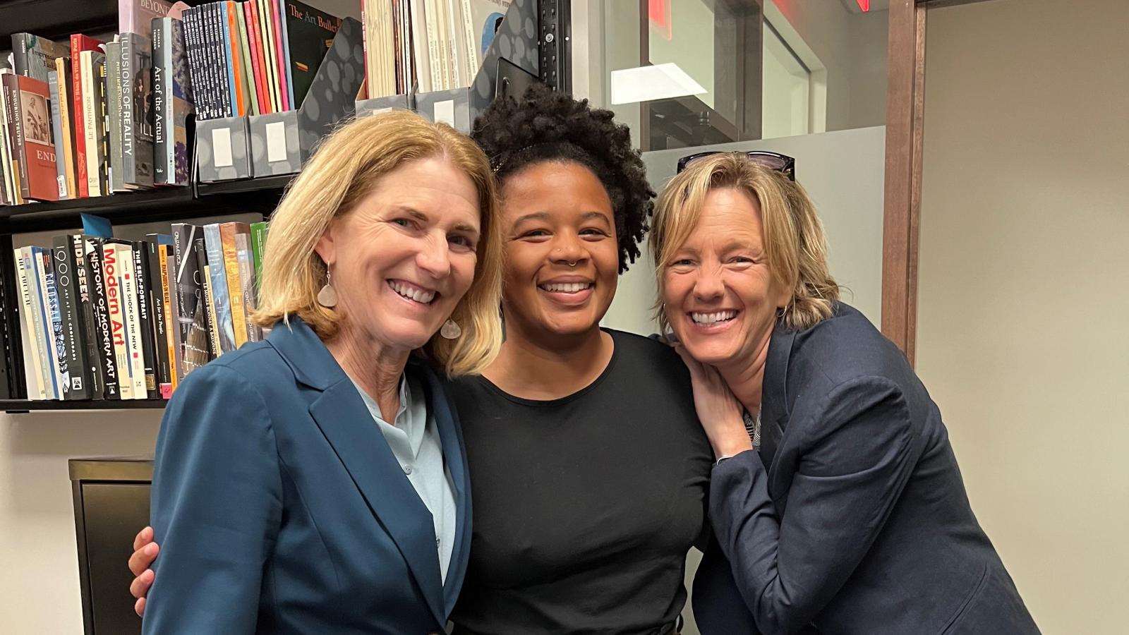 three people standing together in front of a bookcase, smiling