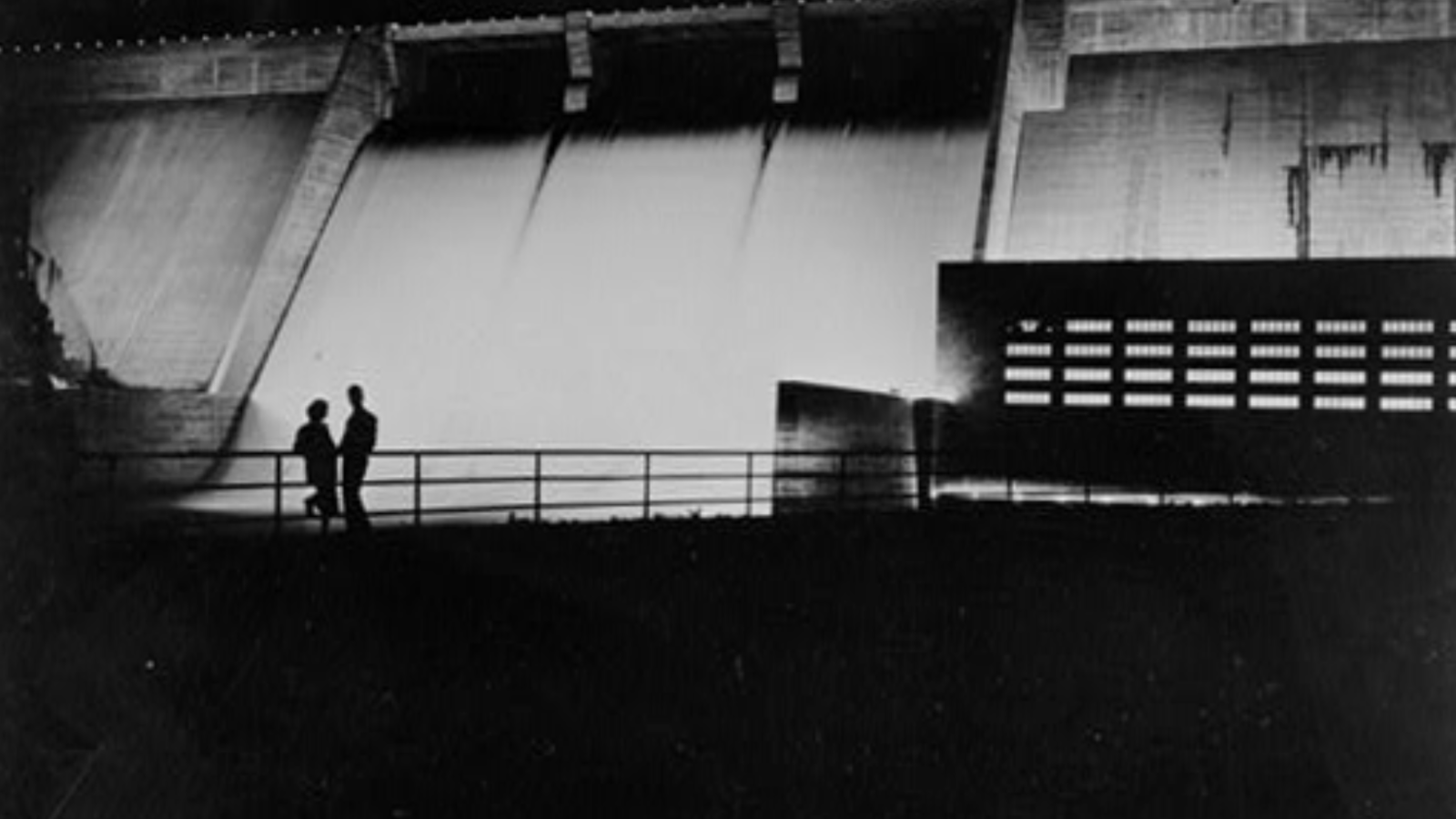 Two people standing by the Norris Dam at night