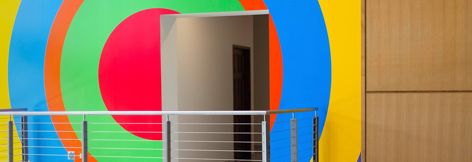 A building interior showing a doorway and balcony with a colorful, geometric mural by the artist Sol LeWitt painted on the wall behind.